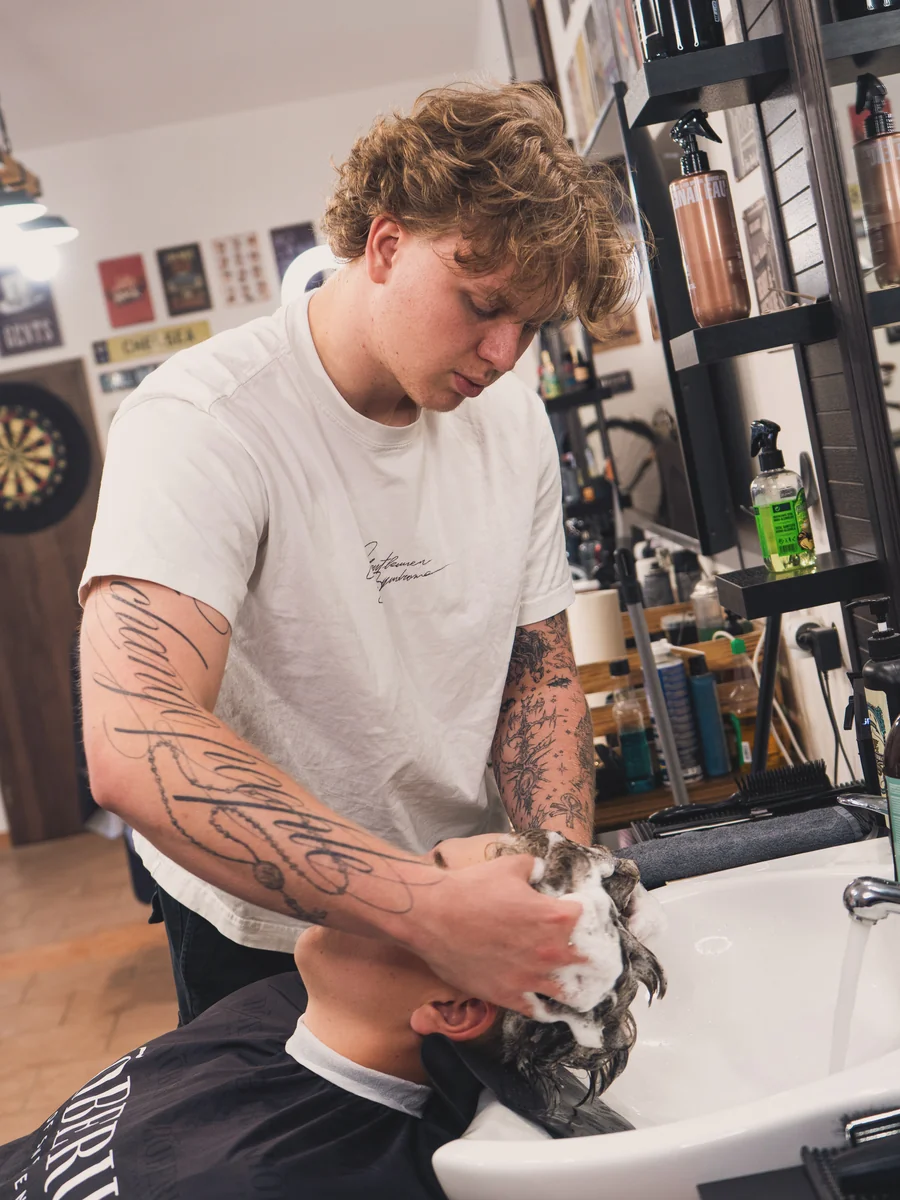 Barbér s tetováním myje vlasy klientovi – Notorious Barbershop Kyjov